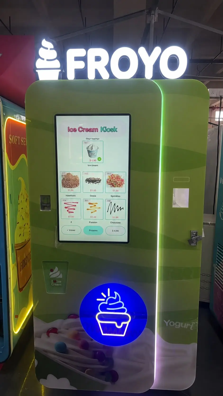 Ein Automat desselben Modells, ganzes Gerät: grünes Gehäuse mit hochformatigem Touchscreen, darüber die Leuchtschrift des Kunden, davor die Becherausgabe.